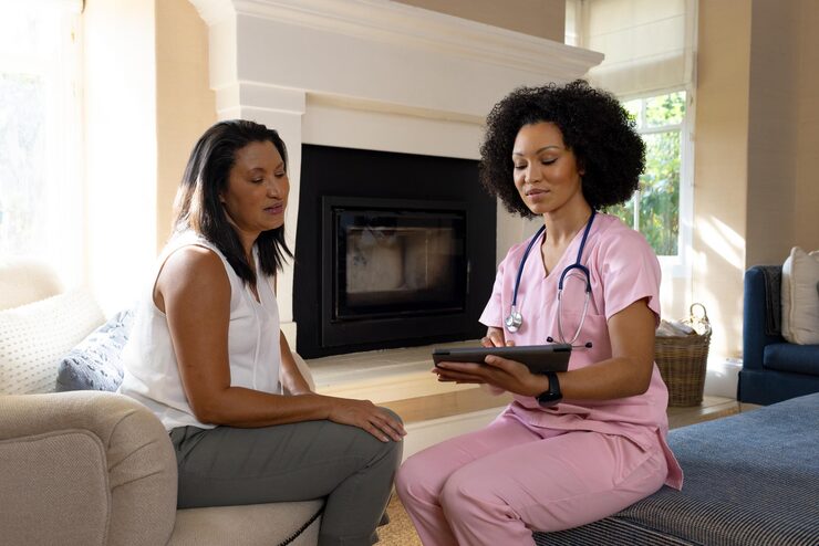 mature biracial woman sitting with young biracial healthcare worker holding tablet home 13339 363174